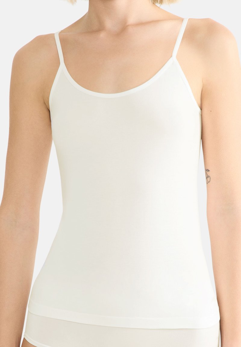 Femme portant un caraco blanc ajusté à fines bretelles, buste supérieur et bras partiels visibles sur un fond uni.