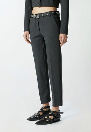 Kvinde iført mørkegrå skræddersyede bukser, en matchende cropped top, et sort bælte med sølvøjer og sorte spidse flade sandaler med stropper.