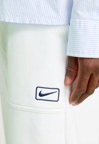 Nærbillede af en hånd, der hviler mod hvide bukser med et marineblåt Nike-logo, kombineret med en lyseblå og hvid stribet skjorteærme.