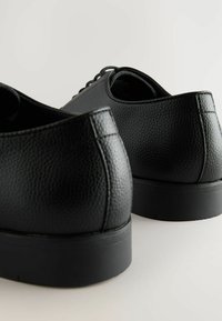 Zapatos de cuero negros con un acabado texturizado, punta redonda y suela de goma negra, que presentan un diseño minimalista sin embellecimientos visibles.