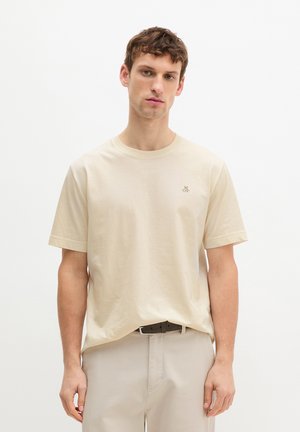 Homme portant un t-shirt à manches courtes beige clair avec un petit logo sur la poitrine et un pantalon beige assorti, debout devant un fond blanc uni.