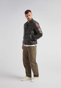 Giubbotto imbottito nero con una toppa rossa sulla spalla, chiusura a zip sul davanti e polsini a costine. Indossato sopra una maglietta bianca e abbinato a pantaloni color khaki e scarpe bianche.