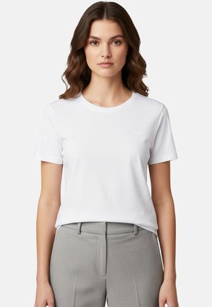 Donna con capelli castani ondulati che indossa una maglietta bianca a maniche corte semplice e pantaloni su misura grigio chiaro su sfondo bianco.