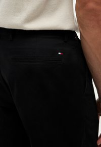 Uomo che indossa pantaloni neri con un piccolo logo rosso, bianco e blu vicino alla tasca posteriore, abbinati a una maglietta di colore chiaro.