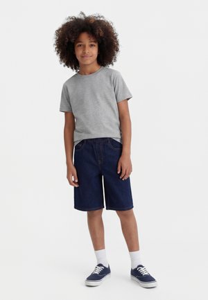 Chlapec s kučeravými vlasmi stojí oblečený v sivej tričku, tmavomodrých rifľových kraťasoch, bielych ponožkách a námornícky modrých teniskách na bielom pozadí.