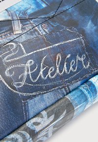 Denimmönstrat tyg med broderi av "Atelier" i vit tråd; har blå nyanser, detaljerad sömnad och texturerad yta.