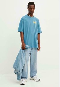 Camiseta de algodón azul con el logo de Vans, combinada con unos jeans de pierna ancha en azul claro. Modelo sosteniendo una chaqueta de mezclilla, usando zapatillas blancas.