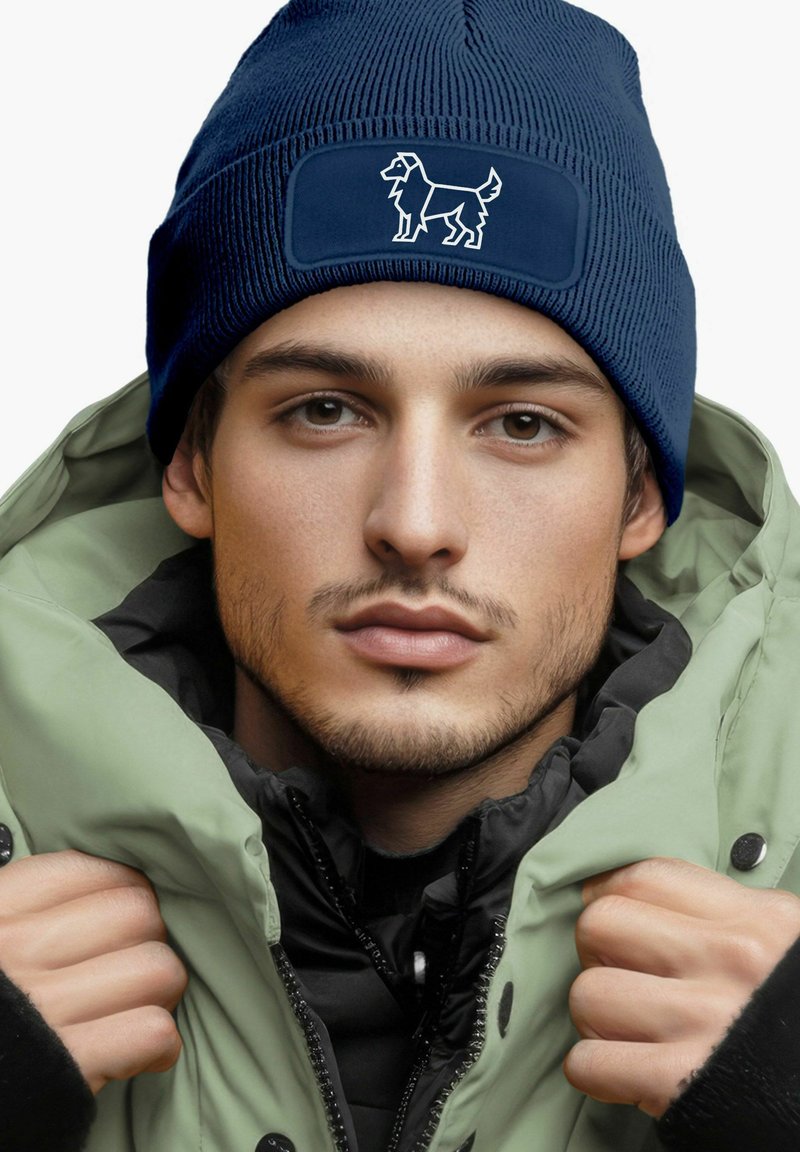 Navy knit beanie with a rectangular patch featuring a white dog silhouette, worn with a light green hooded jacket.