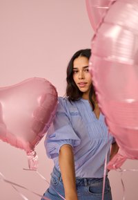 Jeune femme en chemisier bleu clair et jeans tenant des ballons en forme de cœur rose sur un fond rose doux.