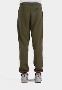 Olijfgroene sweatpants met elastische tailleband en zijzakken; voorzien van contrasterende bruine strepen aan de zijkanten. Gemaakt in combinatie met zilveren sneakers.