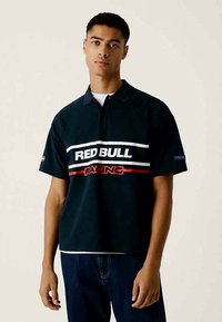 Svart pikétröja med vit och röd "RED BULL RACING"-logotyp. Korte ärmar, klassisk krage och avslappnad passform. Bärs med blå denimjeans.