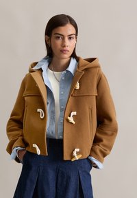 Bruine wolden duffelcoat met een capuchon, voorzien van toggle-sluitingen, gedragen over een lichtblauw gestreept shirt en een donkere spijkerrok.
