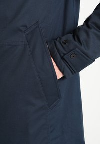 Námořnická modrá látková bunda s detailním pohledem na boční kapsu a zapínací manžetu, s hladkou texturou a dobře padnoucím střihem.