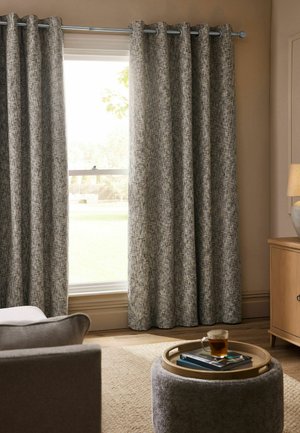 Gray textured curtains partially covering a window in a living room with a gray chair, round ottoman, wooden cabinet, and lamp.