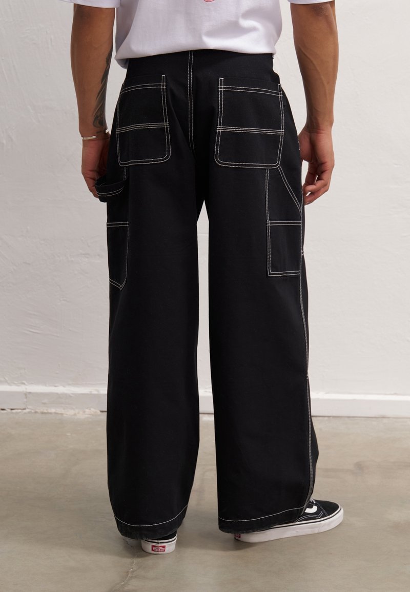 Homme portant un pantalon noir à jambes larges avec des coutures blanches et plusieurs grandes poches à l'arrière et sur les côtés, associé à des baskets noires et blanches.