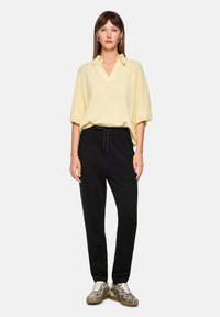 Gelber kurzärmeliger Strickpullover mit V-Ausschnitt, kombiniert mit eng anliegenden schwarzen Hosen mit einem dezenten Zebramuster und Kordelzug in der Taille.