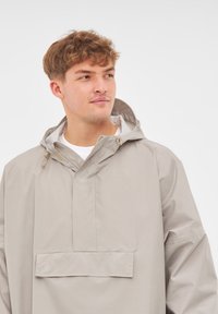 Chaqueta de lluvia con capucha en gris claro, con media cremallera y bolsillo frontal; cuenta con una textura suave y resistente al agua y una capucha ajustable con cordón.