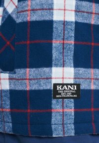 Blå og hvid ternet flanelstof med røde accenter. Har et sort label mærket "KANI THE ORIGINAL EST. 1989" nær kanten.