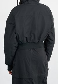 Svart kort jacka med en puffig utsida, elastisk midja och slät yta. Har en hög krage och sydda axelsömmar.