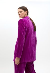 Kvinna med långt brunt hår som bär stora guldörhängen och en strukturerad ljuslila kostym, delvis vänd bort från en neutral bakgrund.