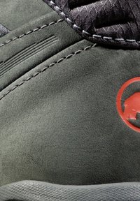 Nærbilde av mørkegrønne semskede vandresko med grå teksturerte paneler, rødt logo og kontraststing. Glatte kanter og robust design.