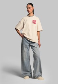 Beige oversize tričko s červeným grafickým textem, spárované s volnými vybledlými modrými džínami a béžovými botami. Stříbrný řetízek a náušnice.