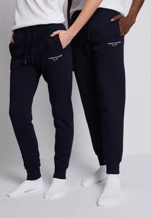 Zwei Personen tragen marineblaue Tommy Hilfiger Jogginghosen mit weißem Logo und stehen nebeneinander mit den Händen in den Taschen vor einem schlichten grauen Hintergrund.