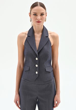 Donna che indossa un blazer senza maniche grigio a righe con tre bottoni e pantaloni abbinati su uno sfondo bianco uniforme.