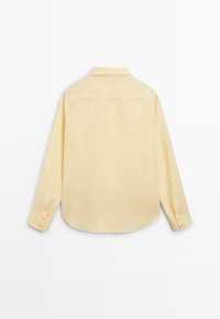 Chemise jaune clair à manches longues avec poignets boutonnés, col à pointes larges et dos uni, posée à plat sur un fond blanc.