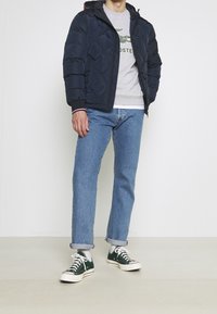 Man i marinblå quiltad jacka, grå sweatshirt med logotyp, ljusblå jeans uppvikta vid anklarna, vita strumpor och mörkgröna låga sneakers.