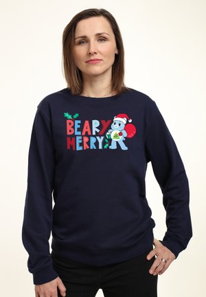 Kobieta w granatowym swetrze z napisem „Beary Merry” i rysunkiem misia w czapce Mikołaja trzymającego ozdobę świąteczną i czerwony worek, stojąca z ręką na biodrze.