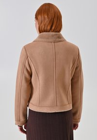 Giacca in faux suede color tan con un morbido colletto in peluche, vestibilità aderente e dettagli delle cuciture. La parte posteriore presenta un design cropped e una texture liscia.