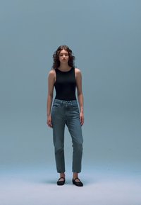 Must värvituumrüü, helesinised kõrge vöökohaga teksad ja mustad libisemisjalatsid; modell seisab helel sinisel taustal.