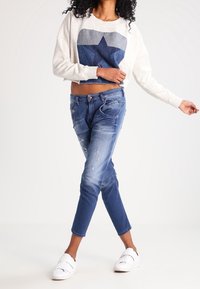 Ljusgrå kortkortad tröja med en mörkblå stjärngrafik, kombinerad med slitna blå denimjeans och vita slip-on-skor.