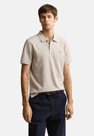 Jeune homme portant un polo à manches courtes beige clair rentré dans un pantalon bleu marine avec une ceinture marron, debout devant un fond uni.