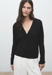 Black cardigan made of soft fabric, featuring a deep V-neck, long sleeves, and button front. Paired with white trousers, minimal design.