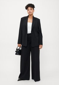 Blazer noir ajusté avec de larges revers, associé à un pantalon noir taille haute. Le mannequin tient un petit sac à main noir. Top blanc en dessous.