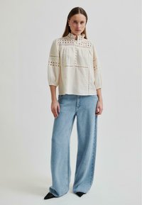 Eine Frau trägt eine cremefarbene, langärmelige Bluse aus Broderie Anglaise mit Knöpfen, kombiniert mit weit geschnittenen hellblauen Jeans und schwarzen spitzen Schuhen.
