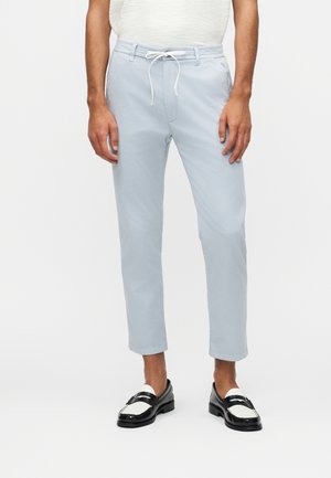 Mężczyzna ubrany w jasnoniebieskie spodnie 7/8 ze białym sznurkiem i czarno-białe mokasyny, stojący na jednolitym tle.