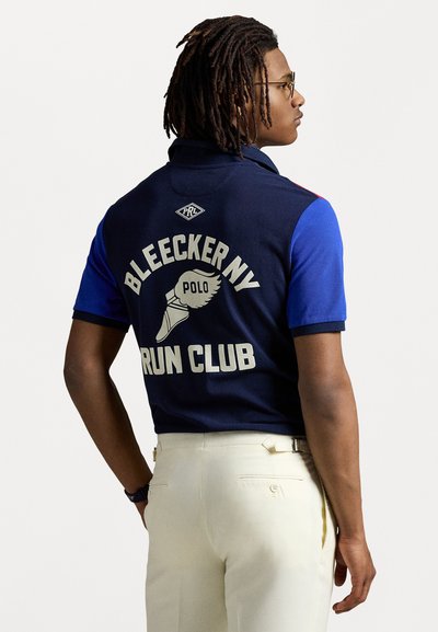 Tengerészkék póló, hátulján a "BLEECKERNY RUN CLUB" felirattal és egy szárnyas lábbal, krémszínű nadrággal párosítva.