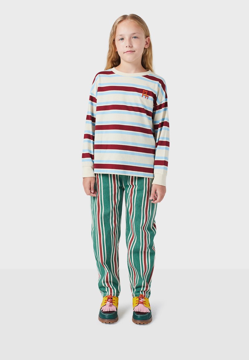 Camisa de manga larga a rayas en azul, blanco y rojo con un pequeño logo. Pantalones de pana verde con rayas verticales en blanco y rojo. Zapatos coloridos.