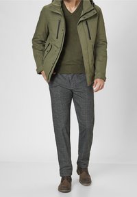 Veste imperméable vert olive avec poches zippées, associée à un pull col en V vert foncé et un pantalon à carreaux gris. Chaussures marron à lacets.