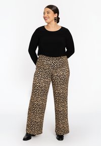 Glanzende vrouw met donker haar in een knot draagt een zwart langetermijn top, broek met luipaardprint en wijde pijpen, zwarte laarzen en zwarte oorbellen, staand tegen een witte achtergrond.