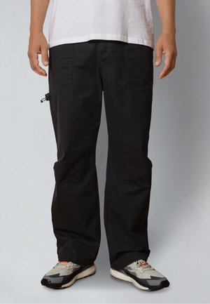 Person wearing black cargo pants with front pockets, standing with hands relaxed, paired with gray and beige sneakers on gray background.