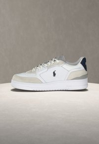 Λευκό και μπεζ χαμηλό sneaker με λευκή σόλα, κορδόνια μπροστά και μικρό μαύρο λογότυπο παίκτη πόλο στο πλάι.