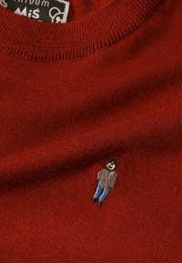 Pull en maille rouge avec une petite figure brodée portant une veste marron et un jean bleu. La texture est lisse avec un col côtelé.