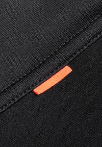 Tessuto nero con una superficie liscia, caratterizzato da un'etichetta arancione prominente cucita lungo la cucitura. Linee pulite e design minimalista.