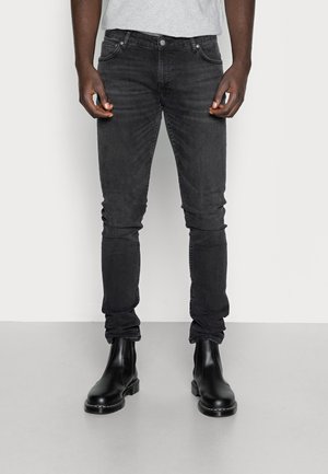 Homme portant un jean noir délavé slim-fit et des bottines en cuir noir, debout devant un fond uni gris clair.