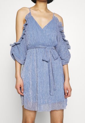 Kobieta ubrana w błyszczącą, jasnoniebieską sukienkę z odsłoniętymi ramionami, cienkimi ramiączkami, falbanianymi rękawami i paskiem wiązanym w talii.