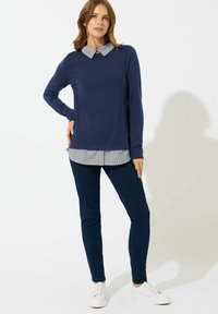 Granatowy sweter z kołnierzykiem, w paski, noszony pod spodem, w zestawieniu z ciemnymi spodniami i białymi sneakersami. Gładka tekstura, casualowy design.